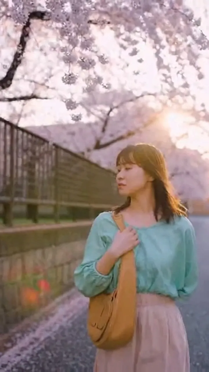 Japanese cinematic realism, poetic spring atmosphere.
A beautiful 21-year-old Japanese female art student walking slowly through a quiet campus lane lined with weeping cherry blossom trees.
Cherry blossom petals drift gently like pale pink snow in the soft spring breeze.
She wears a soft mint blouse and linen midi skirt, natural casual Japanese college student style.
Warm early morning sunlight filters through the blossom canopy, creating soft backlight and subtle lens flare.
The camera slowly follows beside her in a smooth cinematic tracking shot.
Her hair and clothes stir slightly in the wind while petals float around her.
Shallow depth of field, soft muted colors, nostalgic emotional mood, live-action film style.
ultra realistic, cinematic lighting, 4K, film look