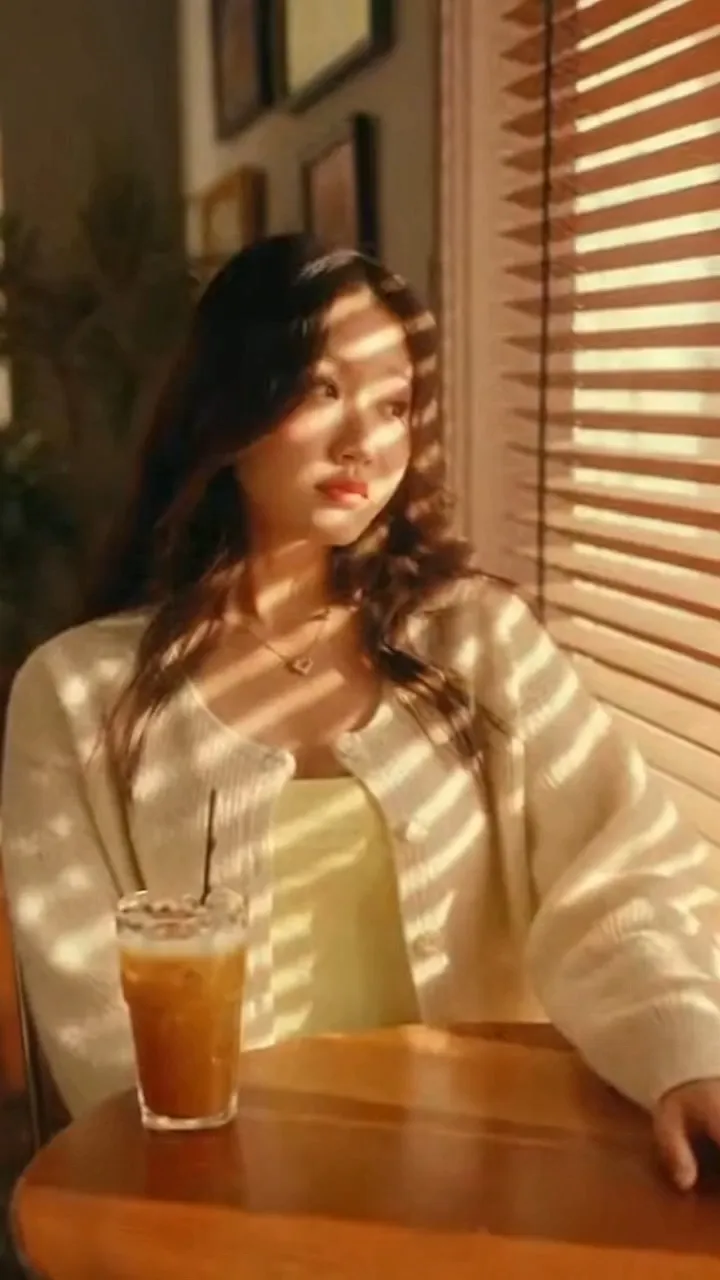 16:9 landscape aspect ratio retro K-pop MV aesthetic warm yellow 35mm film tone nostalgic romantic atmosphere 0-3 seconds opening establishing wide shot vintage 1990s Korean street corner cafe afternoon sunlight streaming through louvered blinds golden dust motes floating in the air camera slowly dollying in a rising young South Korean rookie female idol singer seated at the window side seat soft full baby face gentle drooping willow-shaped eyebrows downturned eyes with thick fluffy lashes prominent dewy aegyo-sal